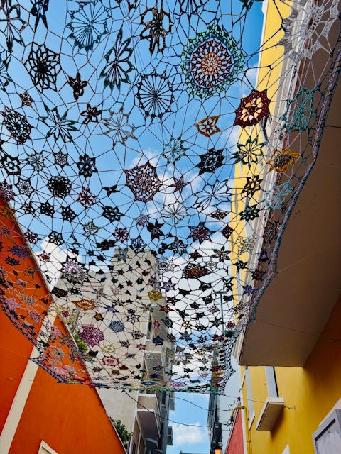 Cielo azul decorado con coloridos encajes de crochet entre edificios amarillos y naranjas. Ambiente alegre y artístico.