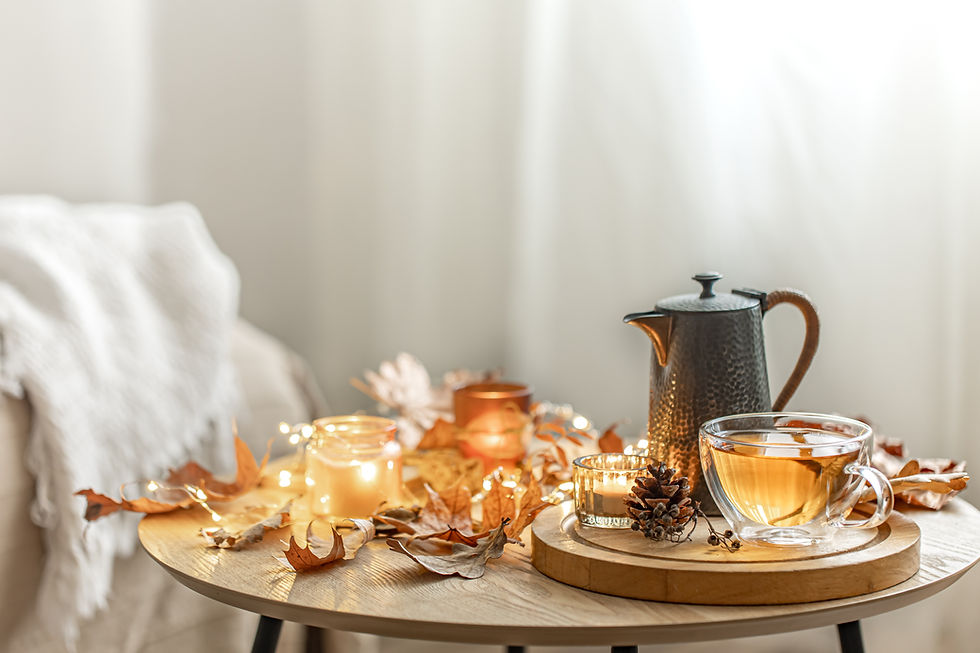 Glas Teetasse mit hellem Kräutertee auf einem Holztisch, daneben eine Teekanne, Kerzen, Lichterkette und Herbstblätter – gemütliche Herbstatmosphäre.