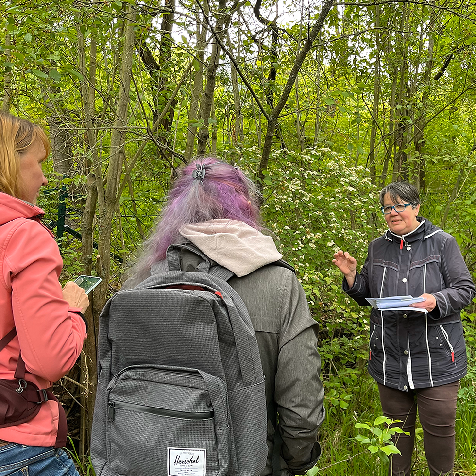 Astrid Hasselmann spricht mit Teilnehmerinnen einer Wildkräuterführung zwischen Sträuchern und Bäume