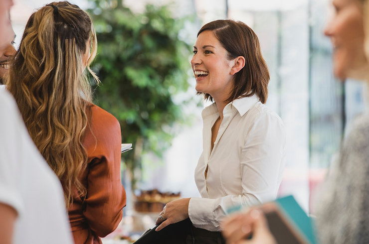 Vrouw in witte blouse lacht tijdens een informele werksituatie met collega’s in een lichte, groene kantooromgeving