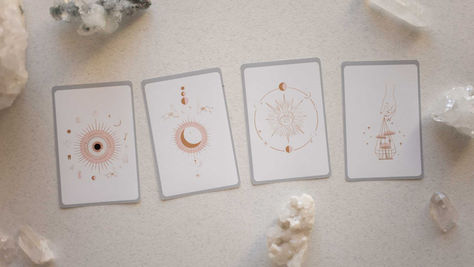 Cartas esotéricas com ilustrações mÃsticas cercadas por cristais sobre uma superfÃcie clara.