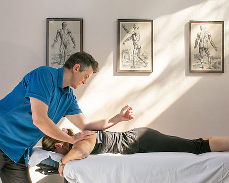 James Tremblay during a Rolfing session with a woman lying on a table. Three anatomical drawings from Andreas Vesalius are on the wall. Soft sunlight fills the room.