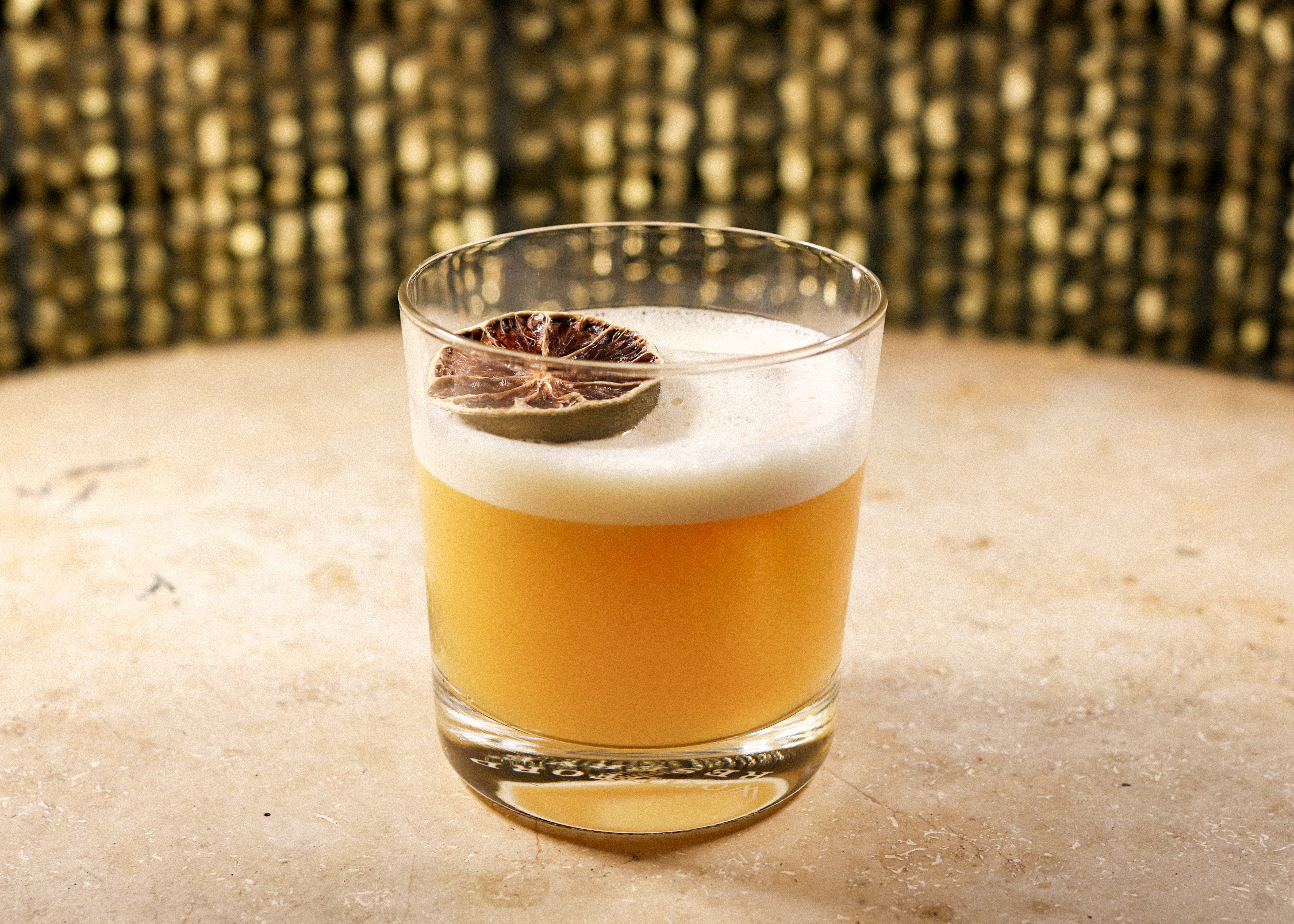 Ein niedriges Glas mit einem orangefarbenen Cocktail und einer weißen Schaumschicht steht auf einer hellen Theke. Auf dem Schaum liegt eine getrocknete Zitrusscheibe. Im Hintergrund ist eine goldfarben schimmernde Wand zu sehen.