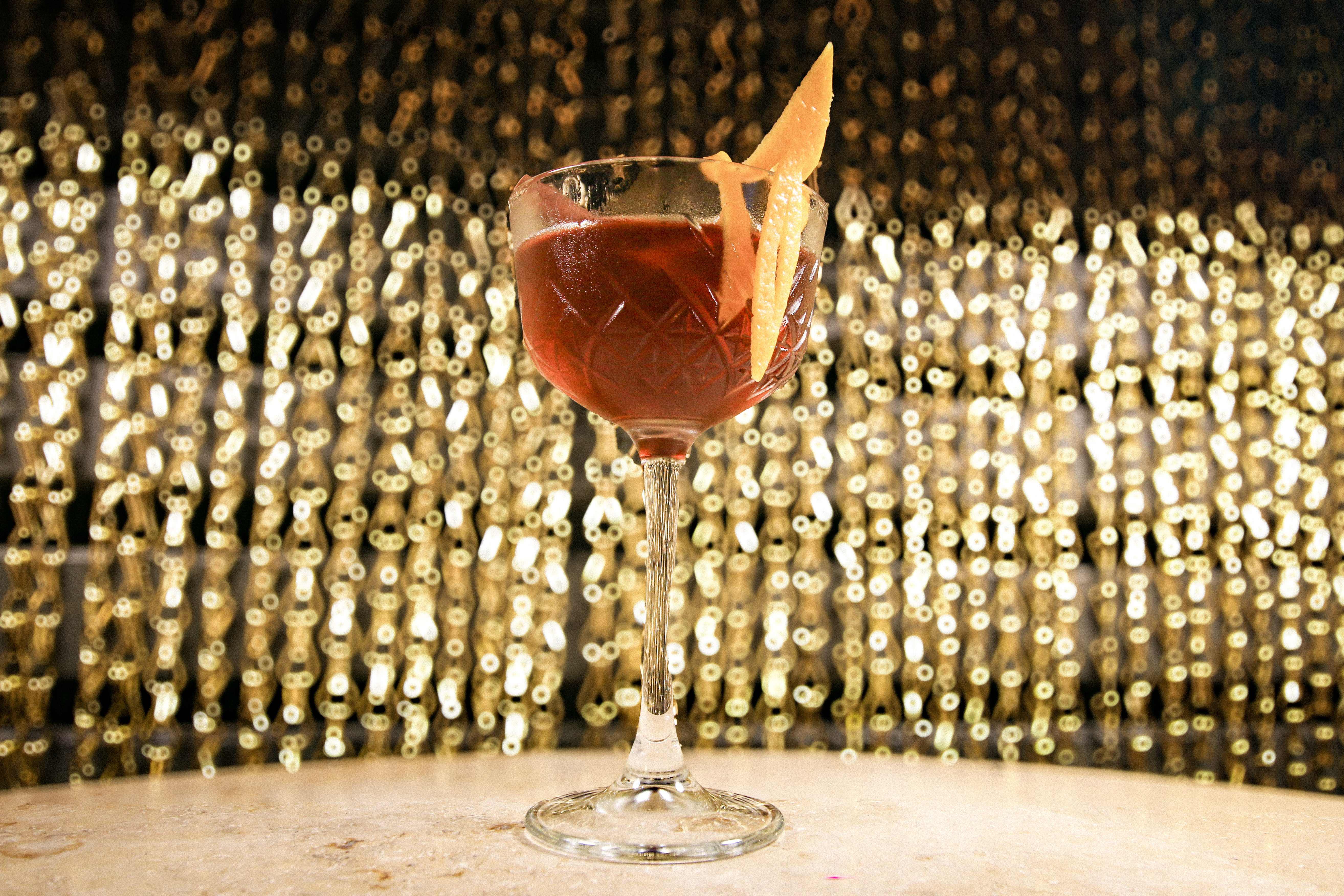 Ein Stielglas mit einem roten Cocktail und einer Orangenscheibe am Glasrand steht auf einer hellen Theke. Im Hintergrund leuchten zahlreiche goldene Lichtpunkte.