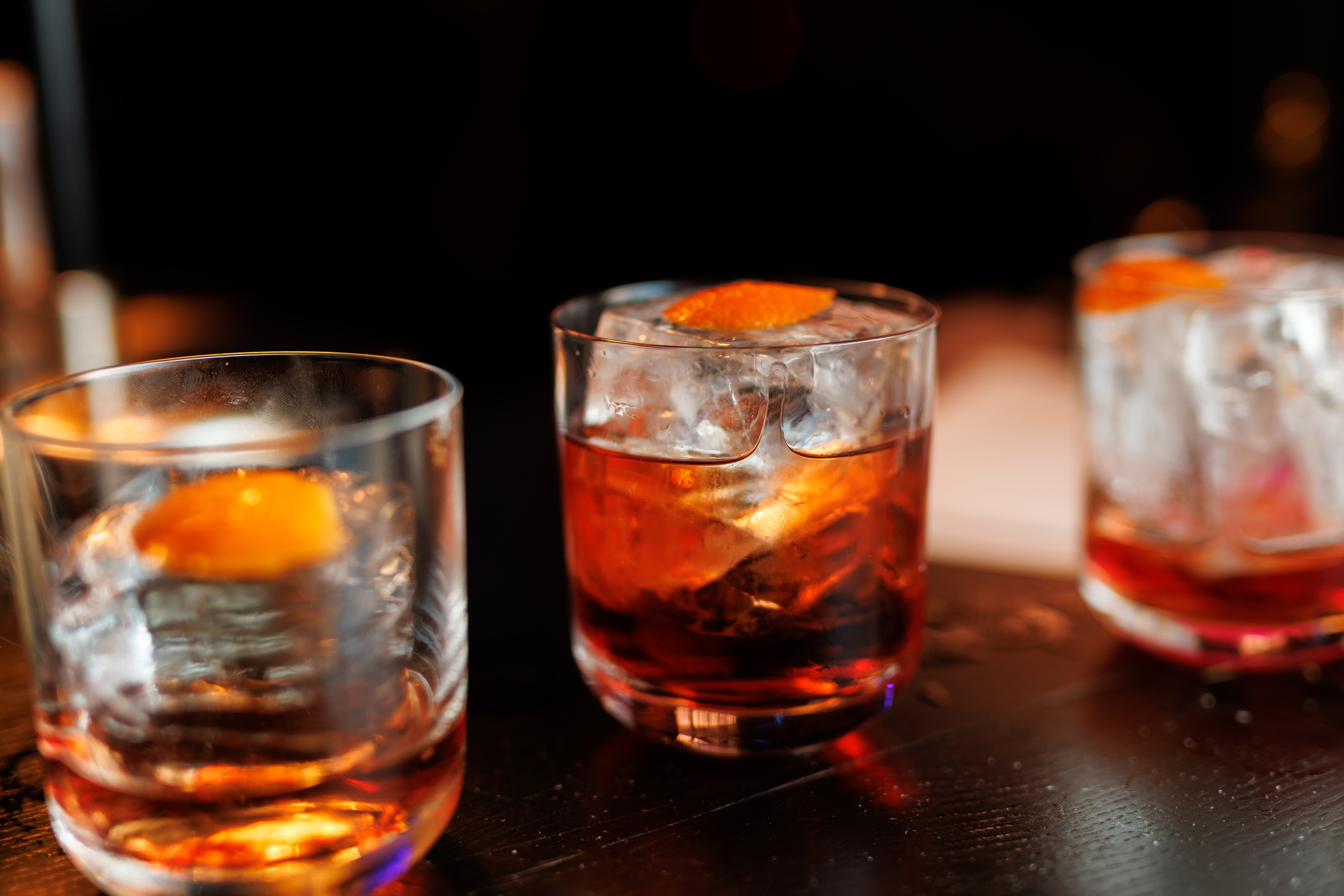 Drei niedrige Cocktailgläser mit rötlichen Getränken, Eiswürfeln und Orangenschalen stehen auf einer dunklen Theke. Das mittlere Glas ist vollständig zu sehen, die beiden äußeren sind angeschnitten. Der Hintergrund ist dunkel und unscharf.