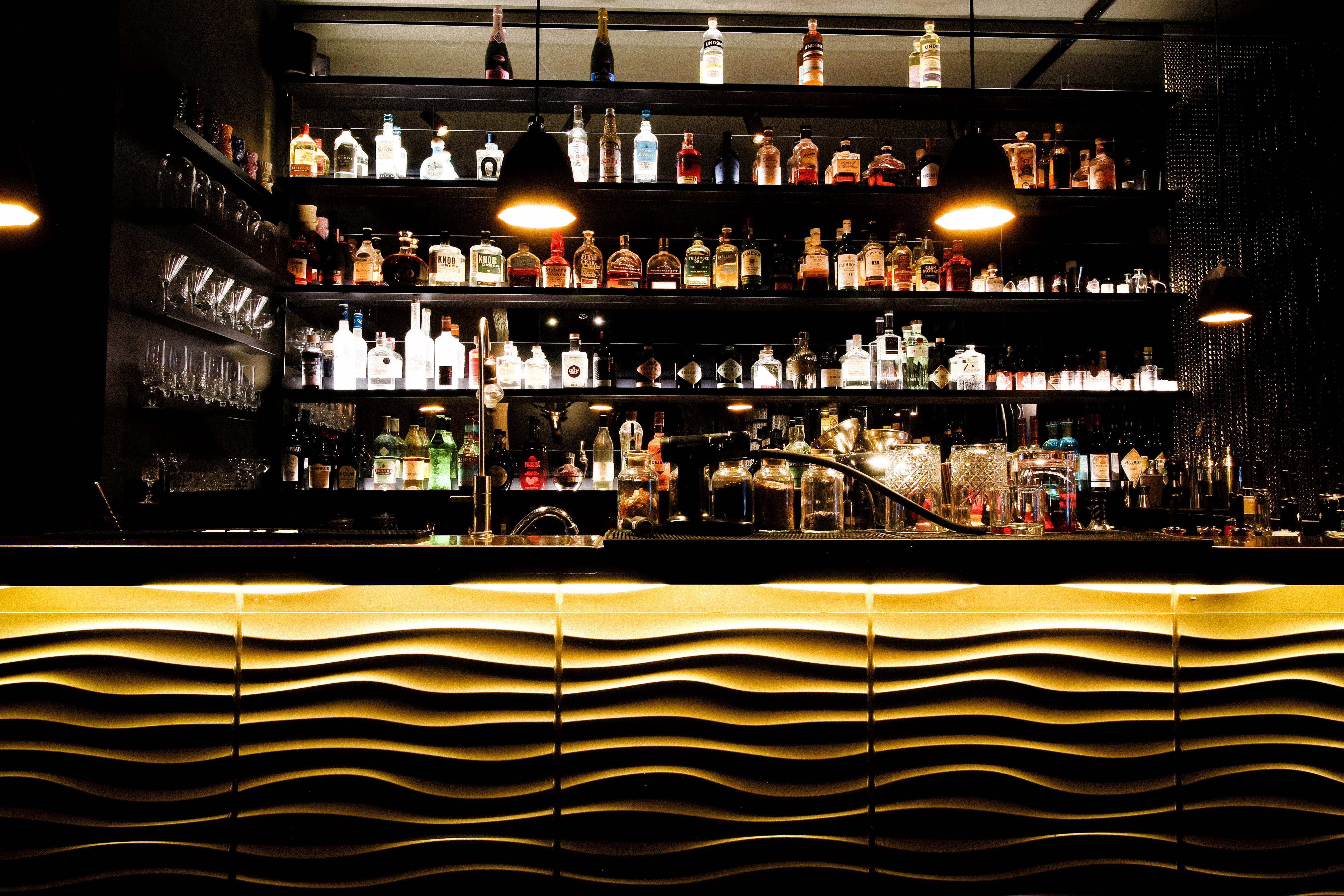 Eine Bar mit mehreren Regalen voller Spirituosenflaschen und Gläser. Zwei warm leuchtende Hängelampen beleuchten den Tresen. Die goldfarbene Tresenfront zeigt ein wellenförmiges Muster.