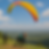 Two people with colorful helmets prepare to paraglide from a grassy hill. A third person watches. The sky is clear, with scattered clouds.