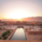 Sunset over a large courtyard with a reflecting pool, surrounded by red buildings and palm trees. People are walking and enjoying the serene view.