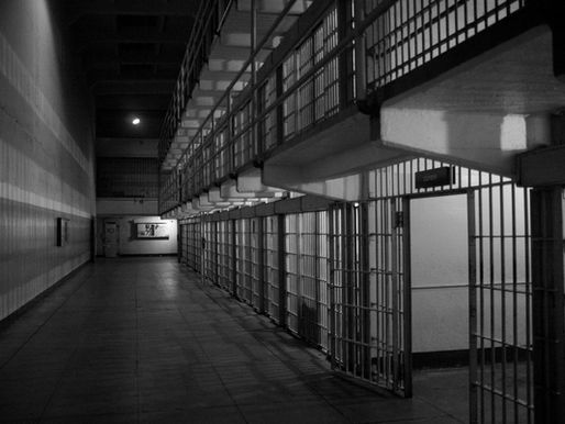 Black-and-white image of an empty prison corridor with barred cells and overhead walkway. Dim lighting creates a somber atmosphere.