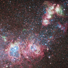 Colorful nebula in deep space, with pink and blue glowing clouds amid dense stars on a dark black background.