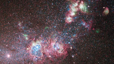 Colorful nebula in deep space, with pink and blue glowing clouds amid dense stars on a dark black background.