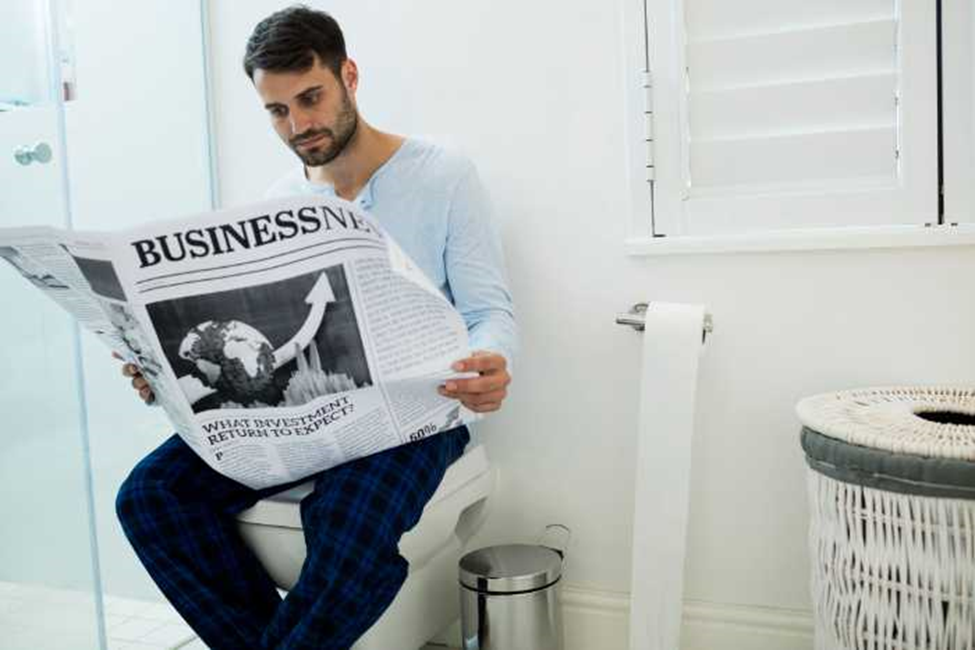 man in bathroom sitting on toilet reading newspaper