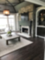 A living room with dark wood floors, a white rug, and a stone fireplace with a dark wooden mantle.