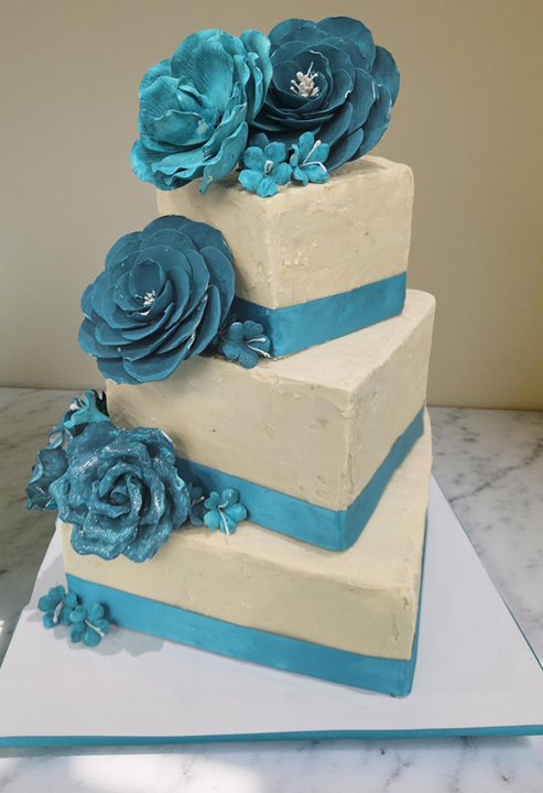 Three-tiered square cake decorated with teal flowers and teal ribbons around each tier, placed on a white square base atop a marble surface.