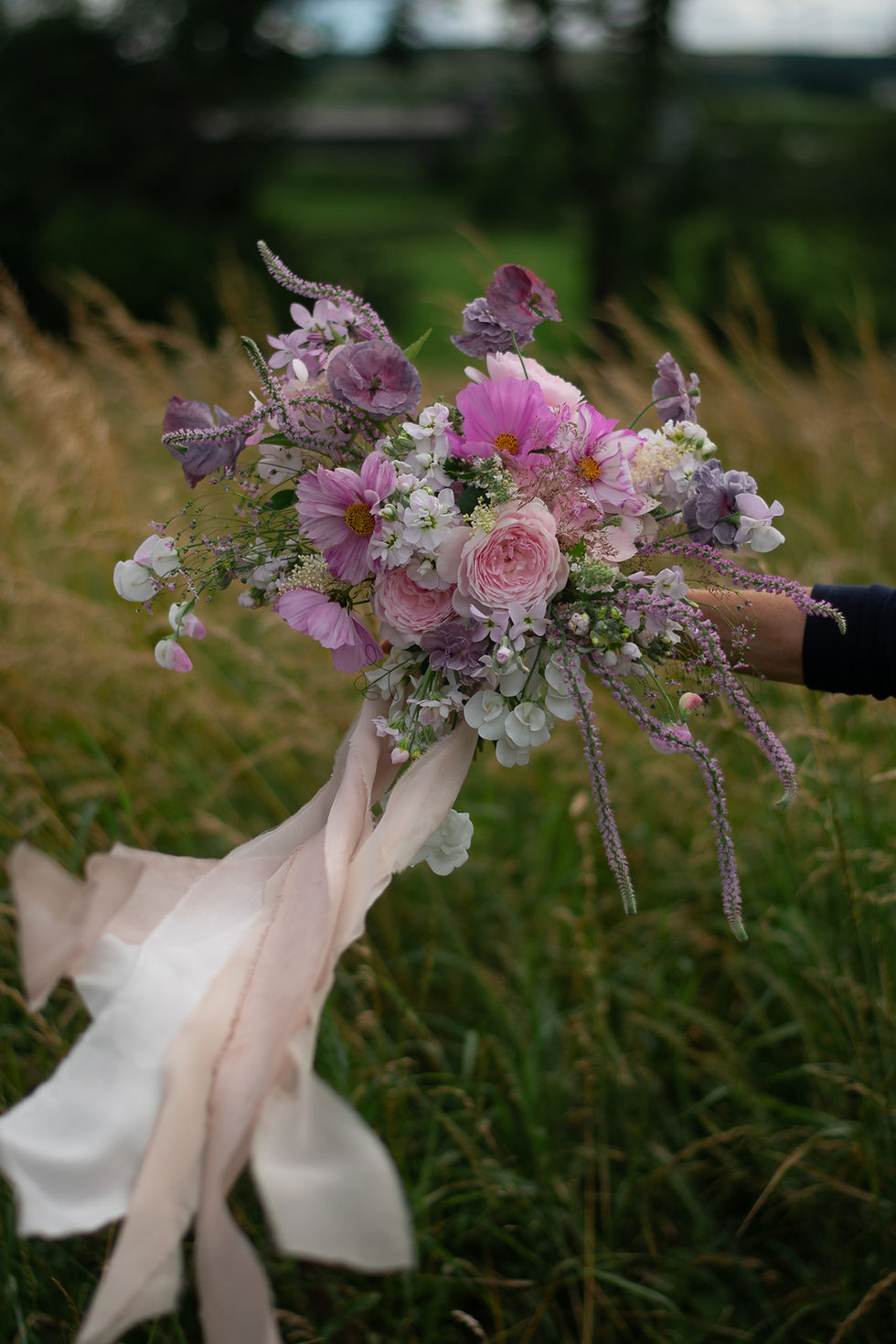 A bridal bouquet of soft blush pink flowers with botanically dyed silk ribbons is held in a hand. Set against a lush green field of golden grasses, creating a serene, romantic mood.