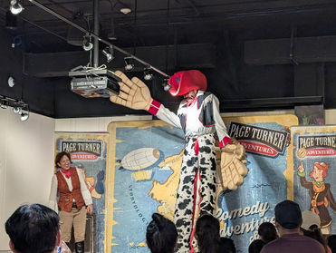 Man on stage in a stilt-walking cowboy costume next to a woman in a steampunk outfit.