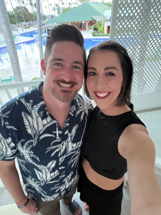 Smiling couple on a balcony overlooking a tropical resort pool.