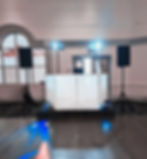DJ booth with speakers and lighting setup in an empty room. Blue and red lights cast on wooden floor. Arched window in the background.