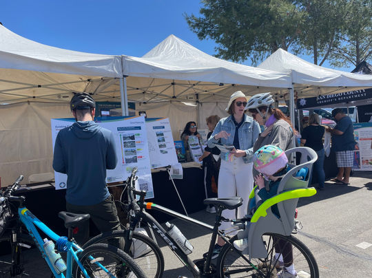 A family with two bicycles (one with a baby carrier, where a baby is sitting with a helmet on their head) stand in front of the interactive map boards. Both adults appear to be reading information about the project, and one is discussing the printed material with a member of the project team. Both adults are still wearing their helmets at the booth.