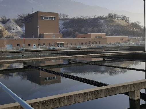 An image of Little Cottonwood Water Treatment Plant shows basins filled with water in front of the brown treatment plant building. In the background are brown and green mountains, with some snow and long brown and green grasses across the landscape.