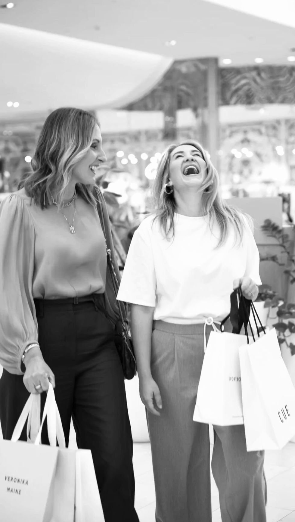 Belinda, Department of Style with client laughing in Karrinyup shopping centre Perth, holding branded bags with "Veronika Maine" and "CUE." Bright and cheerful atmosphere.