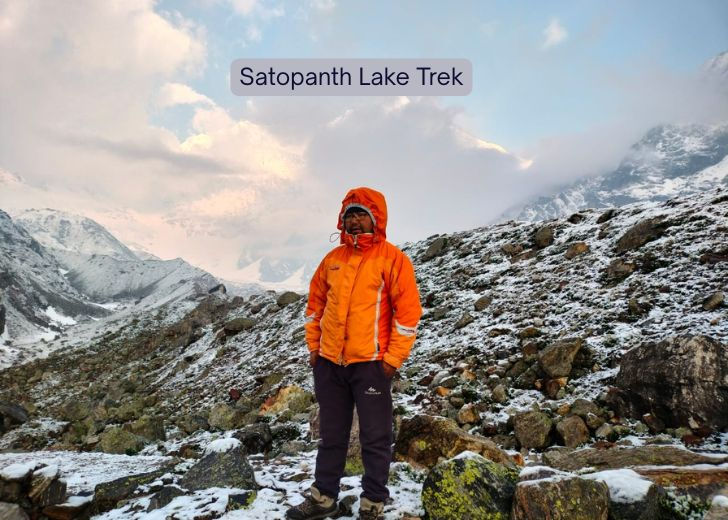 Trekker in orange jacket on snowy slope during Satopanth Lake Trek in Garhwal Himalayas with rugged peaks in background.”