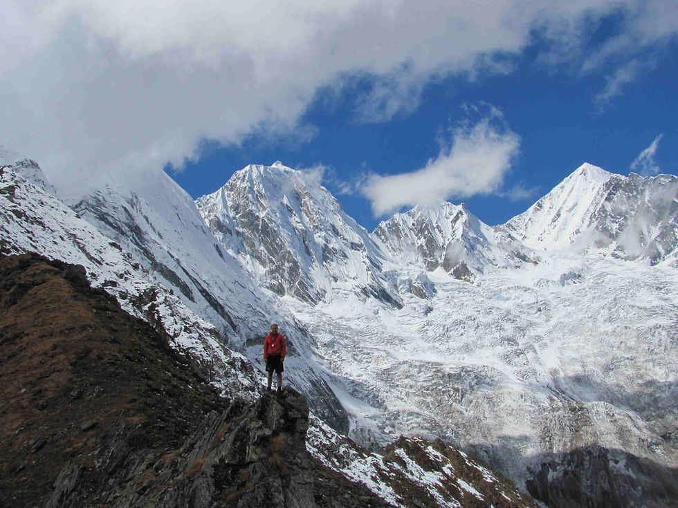 A person in a red jacket stands on a rocky ridge with snow-covered Derma Valley trek in the background. Cloudy sky, sense of adventure.