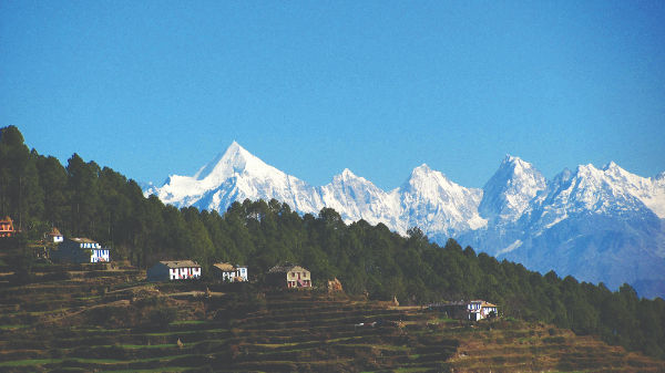 Snow-capped mountains under a clear blue sky, with terraced green hills and scattered houses in the foreground, evoke a serene atmosphere in Kumaon Kingdom.