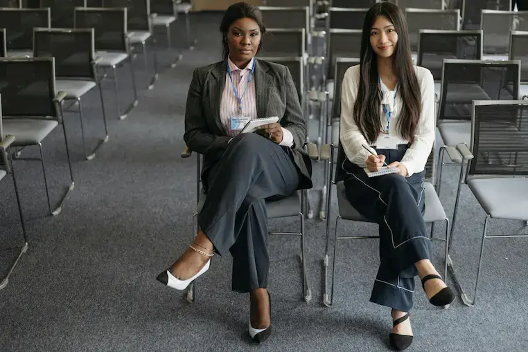 Two women sit in a conference room with note pads, wearing business attire. Rows of empty chairs are visible. The setting feels professional.