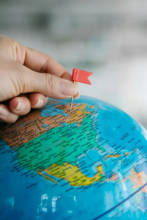 Hand places a red flag pin on a blue globe, focusing on North America. The background is blurred, creating a thoughtful, exploratory mood.