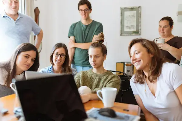 Group of six people gathered around a laptop in a cozy room, smiling and focused. Warm lighting, casual attire, and mugs suggest a relaxed mood.
