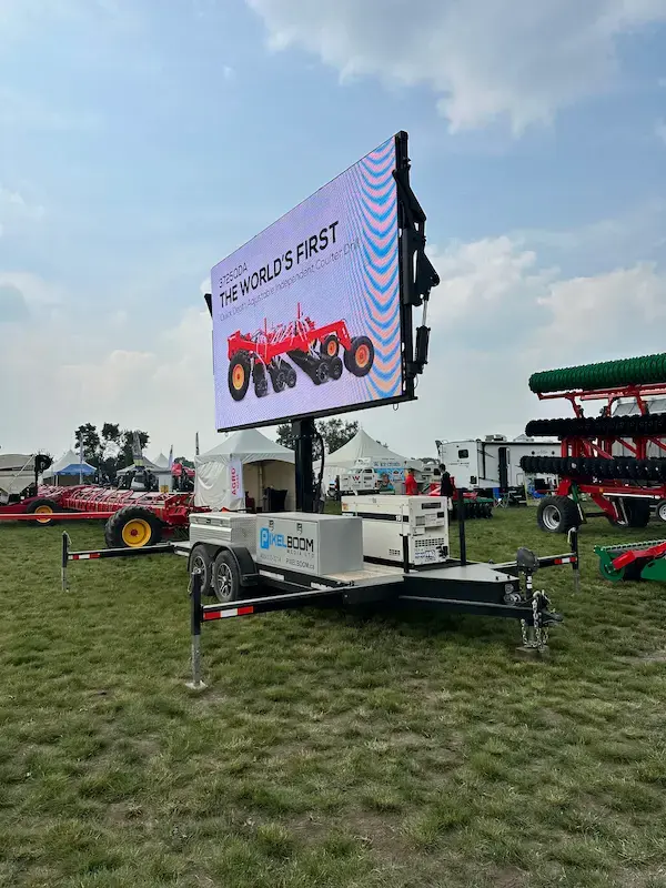 Large screen on grassy field displays red farming equipment ad, reading "THE WORLD'S FIRST." Tents and machinery in background at Ag in Motion.