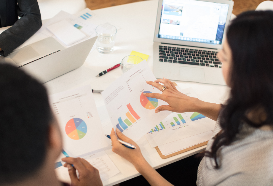 Two ladies at a laptop and looking spreadsheets and chart printouts