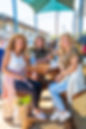 A group of three women sitting at a picnic table at First Monday Canton smiling at the camera.