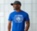 Man in a blue shirt with white text "I'm Coming Home Alumni," smiling and standing against a tiled wall. Wears a black cap, cheerful mood.