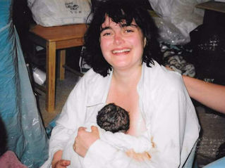 woman smiling, holding newborn baby, in her bedroom, with midwives nearby