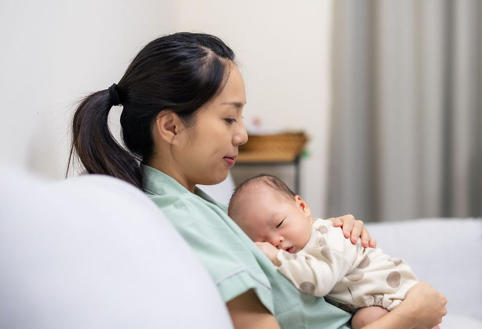 An Asian woman is sitting back on a sofa. She is holding her sleeping baby on her chest. She looks pensive.