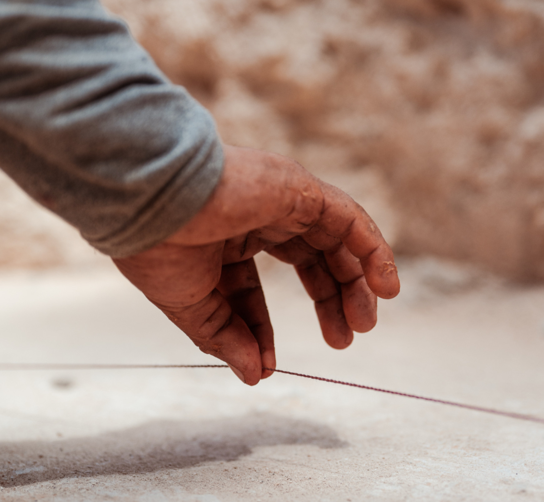 Close-up of a dirty hand in a gray sleeve pulling a thin string over sandy ground, with a blurred tan background.