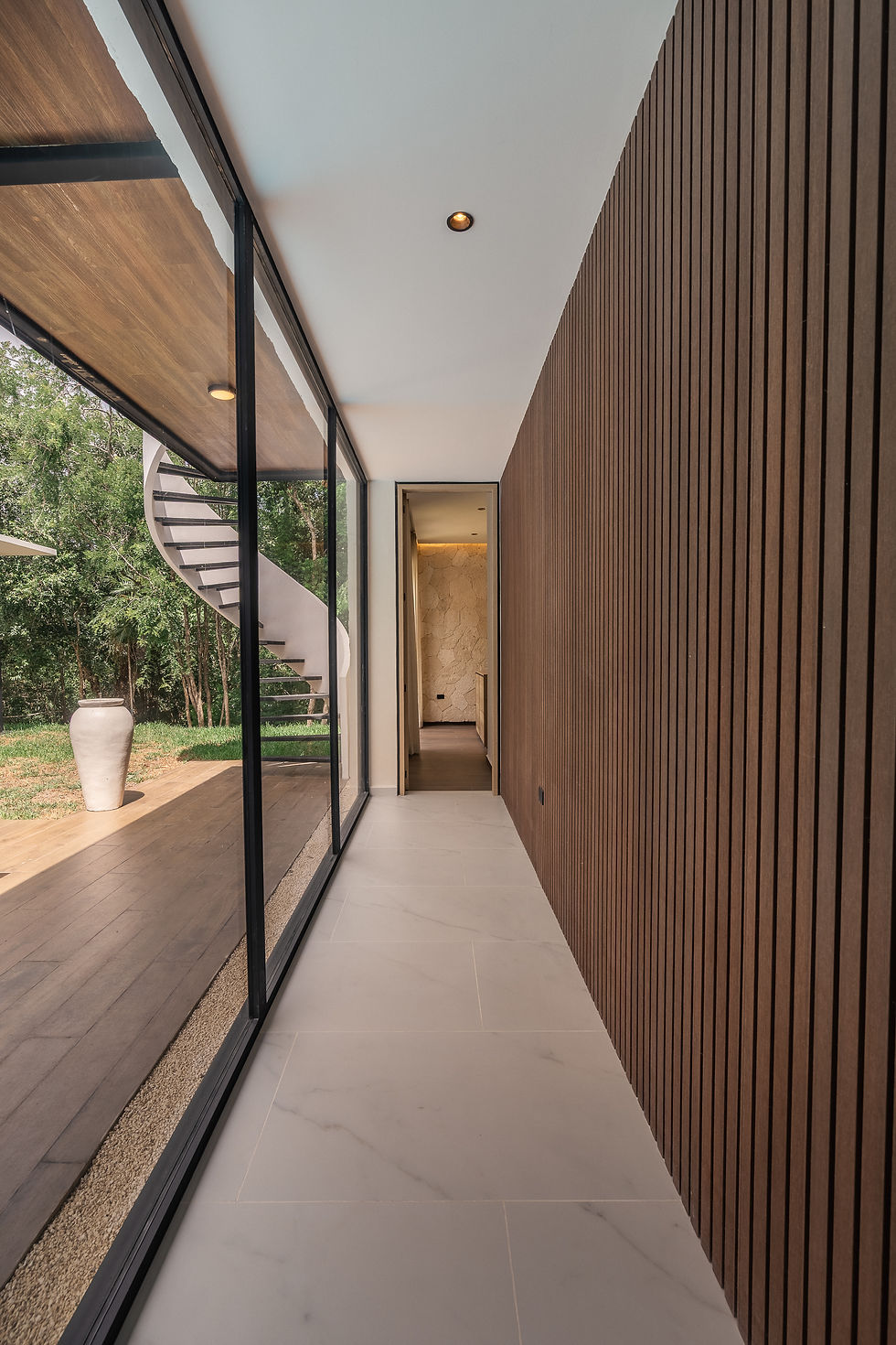 Modern hallway with glass wall, wood slat paneling, spiral staircase outside, and a large white vase beside trees.
