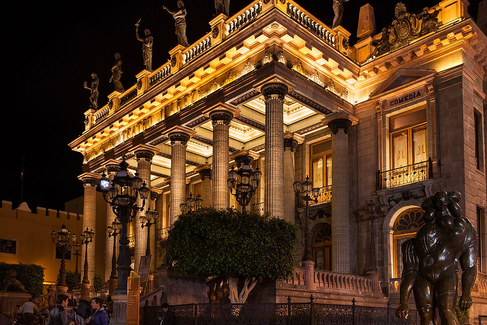 Fachada del Teatro Juárez iluminada por la noche, con columnas y estatuas. Letrero "COMEDIA". Farolas en el frente y personas paseando.