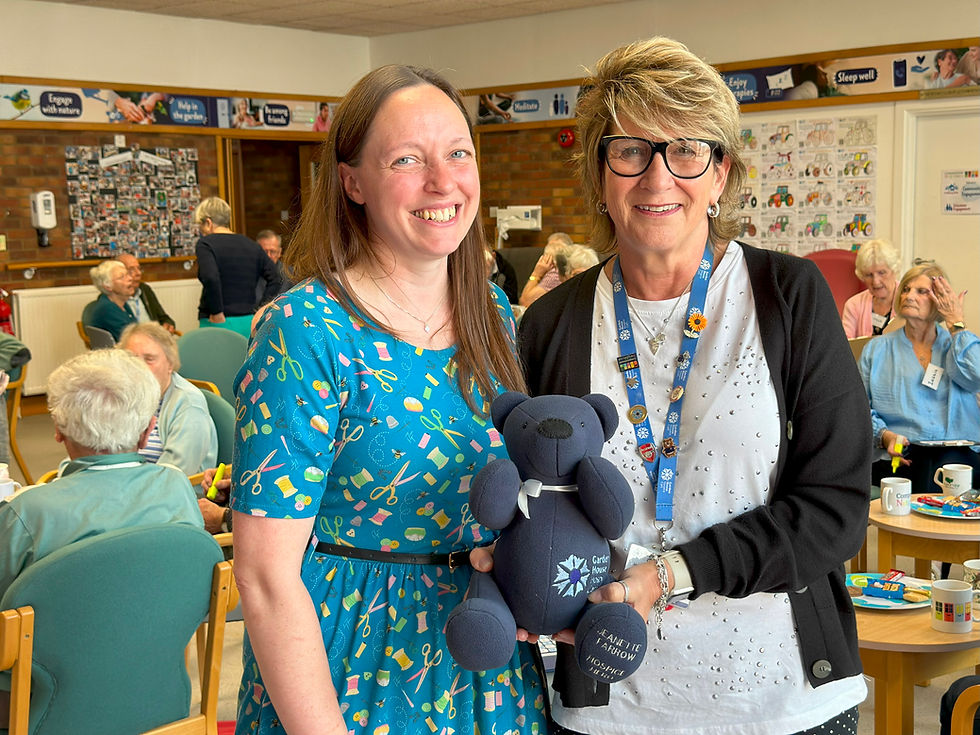 2 women smiling holding a beautiful blue bear. in the background is a number of people enjoying a chat and cups of tea