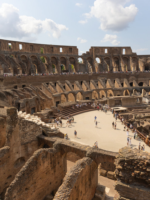 Wide interior view of the Colosseum’s main floor in Rome, showing tourists walking across the ancient stone arena floor beneath the surrounding arched walls and tiered structure of the amphitheatre.