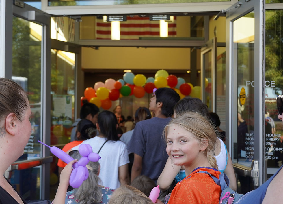 A female student with blonde hair looks back as she enters the doorway of a school, surrounded by multiple people and balloons in the distance.