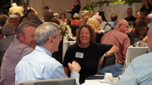 In a crowded room, a woman sits and laughs as two gentleman tell stories.