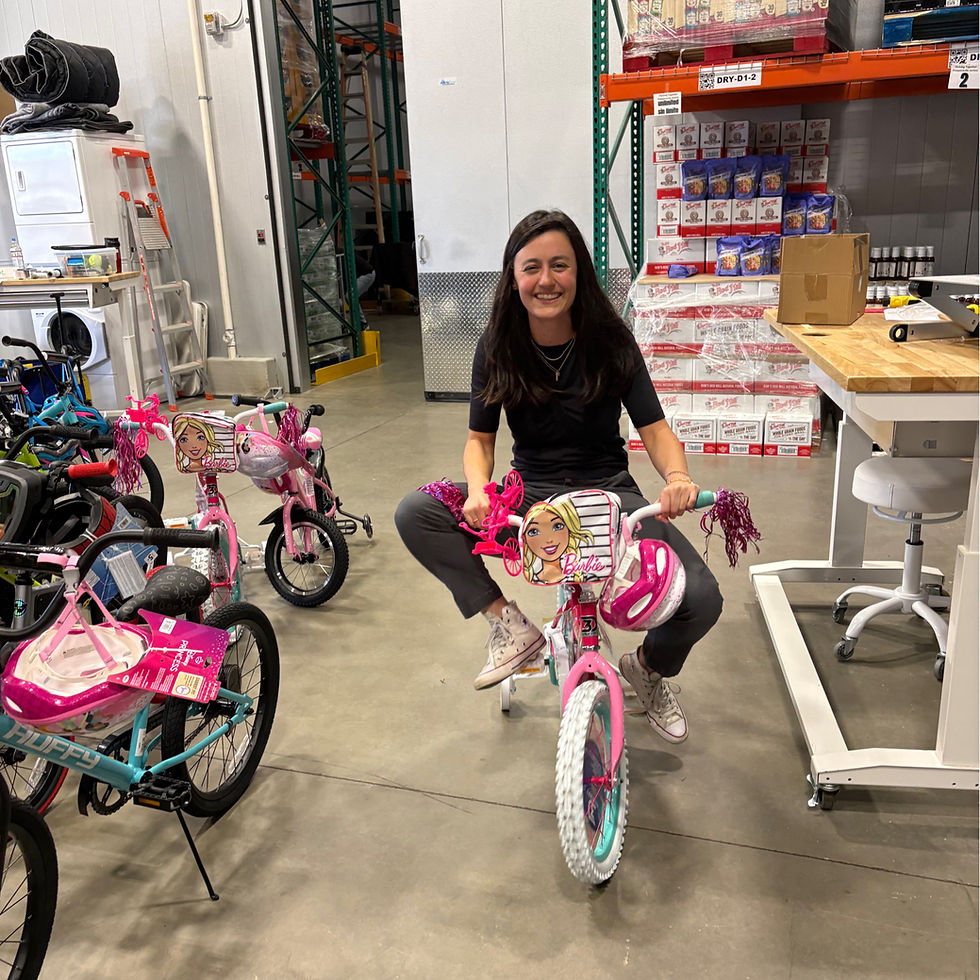 Woman smiling while sitting on a pink Barbie bike in a warehouse filled with packaged goods and other bikes. Playful and cheerful mood.