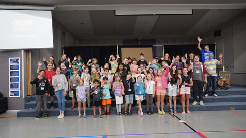 A large group photo of students and volunteers stand on a stage, waving at the camera.