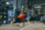 Two people stretch in a gym, raising arms in lunges. They wear bright athletic wear. Gym equipment visible.