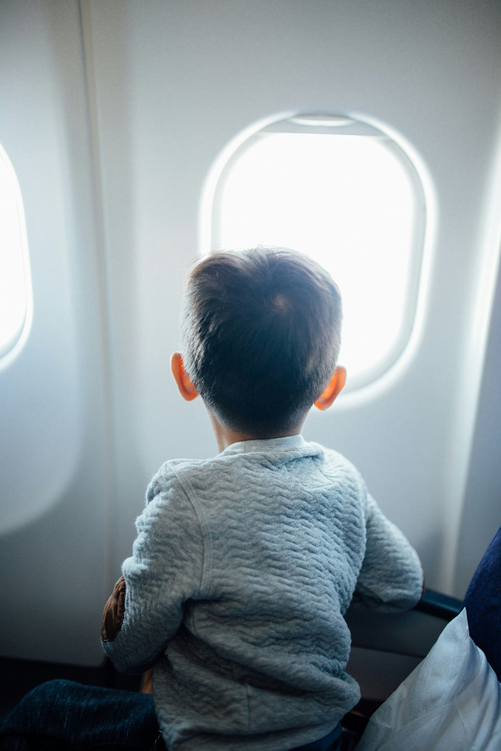Young boy in a gray sweater looks out a bright airplane window, seen from behind, quiet and contemplative.