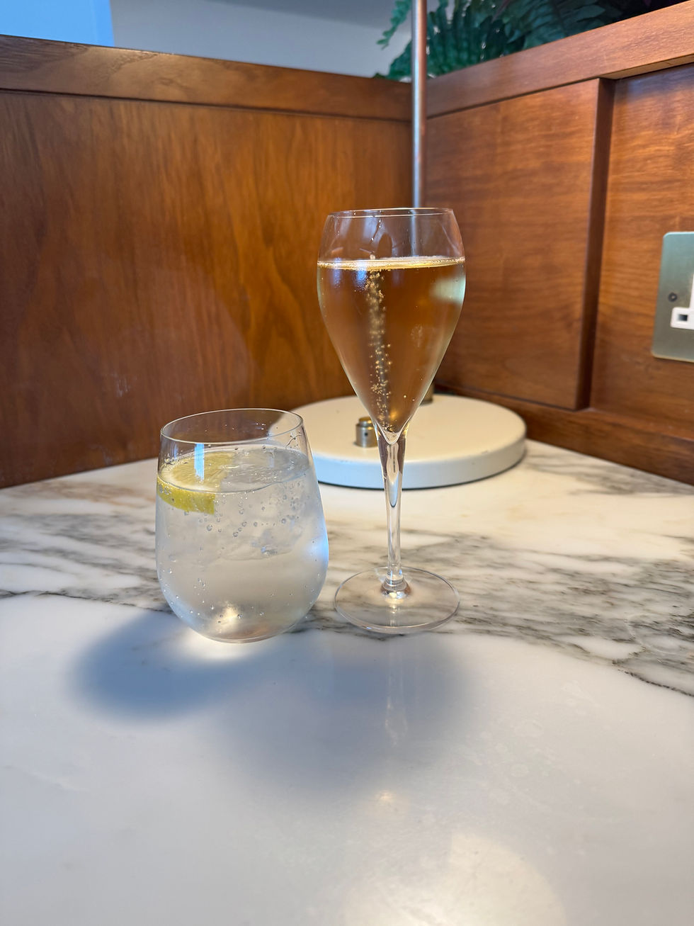 Two glasses on a marble table: one with champagne bubbles and one with a lemon slice in sparkling water. Wooden wall background.
