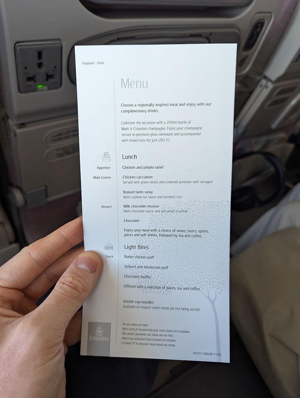Hand holding an Emirates menu on a flight, offering lunch and light bites. Visible seating area in background. Menu text includes food and drink options.
